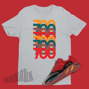 Puede incluir: Una camiseta gris con un gr&aacute;fico apilado del n&uacute;mero 700 en diferentes colores. La camiseta se combina con zapatillas rojas, naranjas y negras.