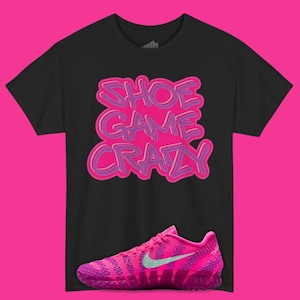 May include: Black t-shirt with the text "SHOE GAME CRAZY" in pink and purple. A pink and purple athletic shoe with a green swoosh is displayed below the shirt. The background is pink.