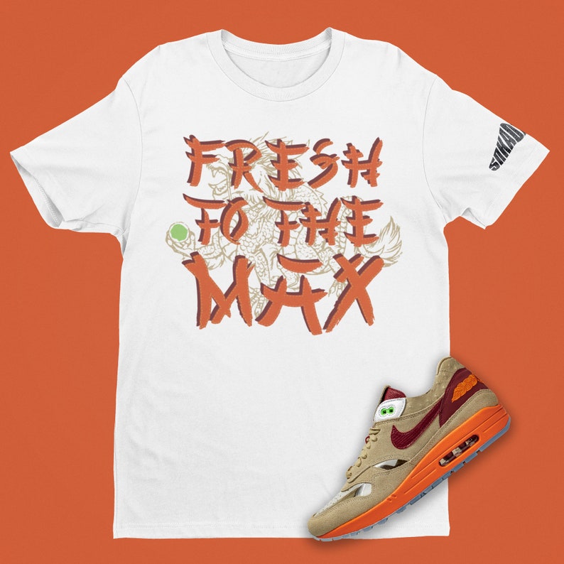 May include: White t-shirt with orange text that says "FRESH TO THE MAX" with a graphic of a dragon. A pair of tan, orange, and red Nike Air Max 1 sneakers are shown below the t-shirt.