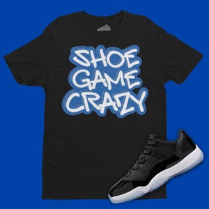May include: Black t-shirt with blue text that says "Shoe Game Crazy". A black and white Air Jordan 11 shoe is on a blue background.