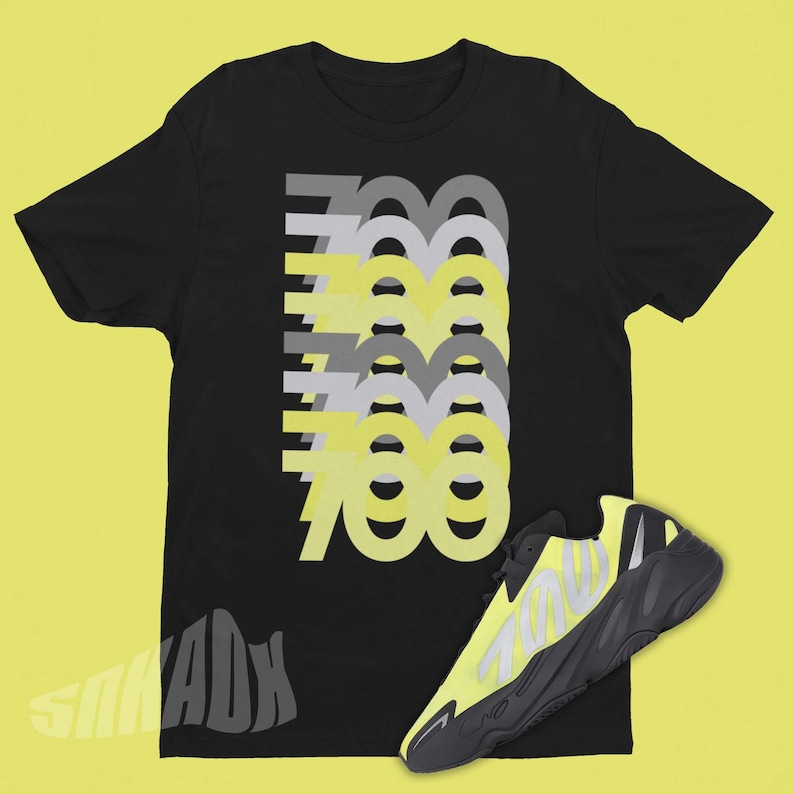 Op de afbeelding: Zwart T-shirt met een afbeelding van het getal 700, herhaald in geel en grijs. Een paar zwarte en gele sneakers met het getal 700 aan de zijkant.
