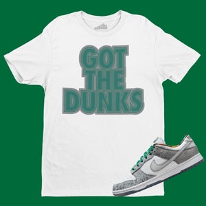 May include: White t-shirt with the text "GOT THE DUNKS" in green and gray lettering. A pair of gray and green Nike sneakers with a white swoosh.