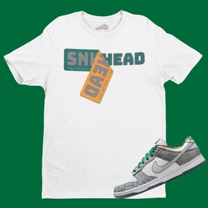 May include: White t-shirt with the text "SNEAKHEAD GOT 'EM ALL" and a tag that says "JUST  GOT 'EM ALL" in grey text on a yellow background. A pair of white, grey, and green sneakers with a white swoosh logo.