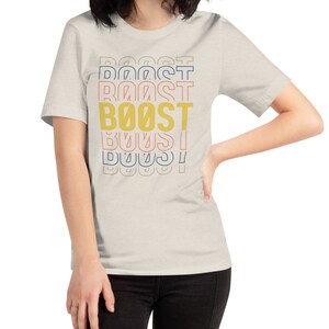 May include: A light beige t-shirt with a graphic print of the word "BOOST" in yellow, with the word "BOOST" repeated in different colors above and below the main word.