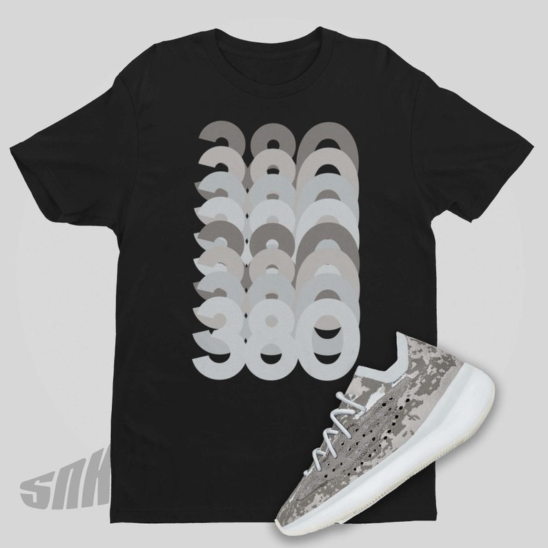 Puede incluir: Una camiseta negra con un gr&aacute;fico del n&uacute;mero 380 repetido en diferentes tonos de gris. Un par de zapatillas blancas y grises con un patr&oacute;n de camuflaje se muestra debajo de la camiseta.