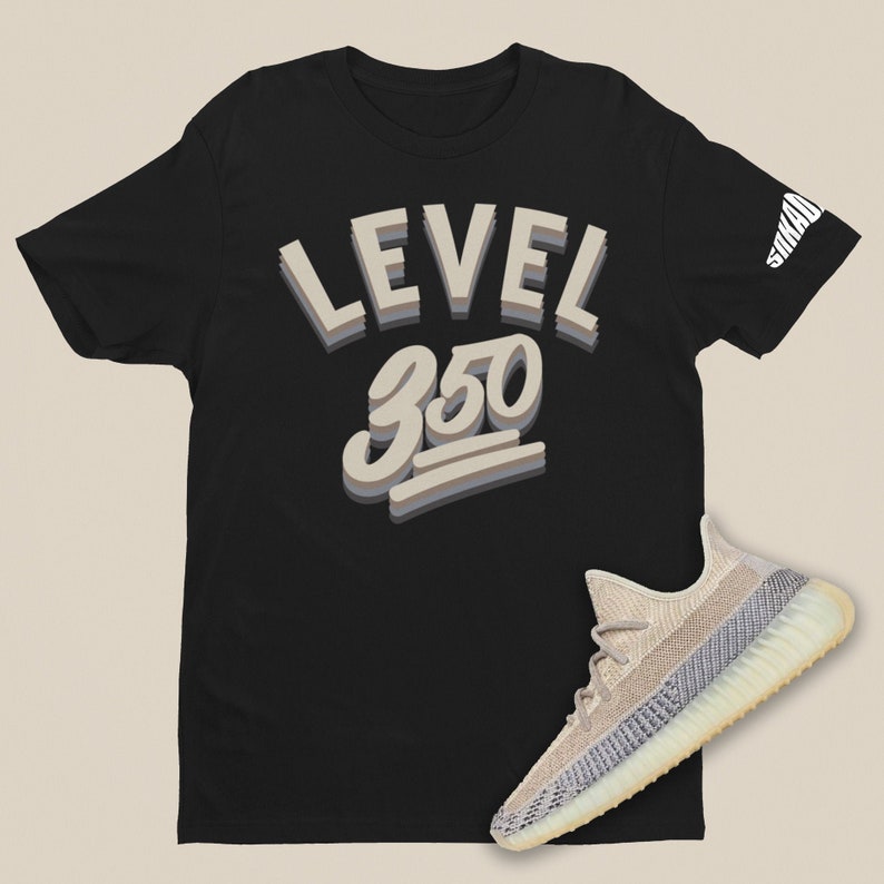 Puede incluir: Camiseta negra con el texto "LEVEL 350" en estilo 3D. Un par de zapatillas beige y blancas se encuentran en la parte inferior derecha de la imagen.