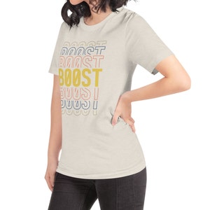 May include: A light beige t-shirt with the word "BOOST" printed in yellow, red, blue, and pink letters. The letters are stacked on top of each other, with the word "BOOST" in the center.