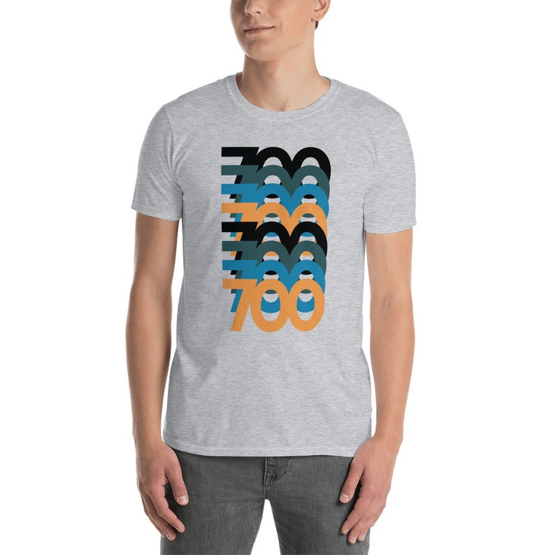 Peut inclure: Un t-shirt gris chin&eacute; avec un motif graphique du nombre 700 r&eacute;p&eacute;t&eacute; dans diff&eacute;rentes couleurs, notamment le noir, le bleu, le bleu sarcelle et l'orange.