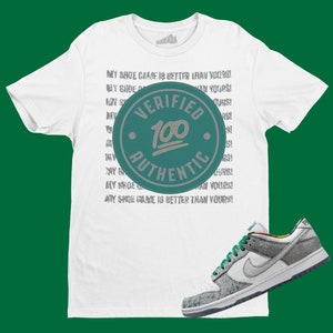May include: White t-shirt with the text "MY SHOE GAME IS BETTER THAN YOURS!" repeated multiple times. A green and grey circular sticker with the text "VERIFIED 100 AUTHENTIC" is on the shirt. A pair of grey and green sneakers are on a white surface.