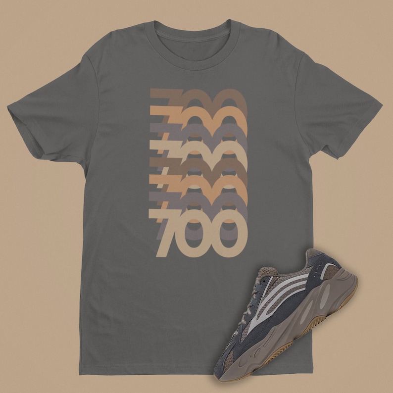 May include: A dark gray t-shirt with a stacked graphic of the number "700" in various shades of brown, beige, and gray. A pair of gray and brown sneakers are shown below the shirt.