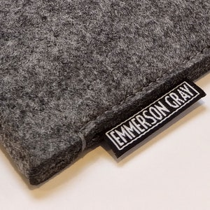 May include: Close-up of a gray felt item with a textured surface. A black rectangular tag with the text "EMMERSON GRAY" in white is attached to the item.