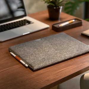 May include: A grey felt laptop sleeve with a white and orange stylus on a wooden desk. A laptop, pen holder, notepad, and small plant are also on the desk. The desk is made of dark wood.