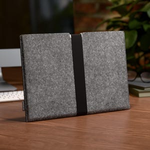 Könnte beinhalten: Eine graue Laptop-Hülle aus Filz mit einem schwarzen Gummibandverschluss. Die Hülle ist rechteckig und steht auf einem Holzschreibtisch. Ein Computermonitor, eine Tastatur und andere Büromaterialien befinden sich im Hintergrund.