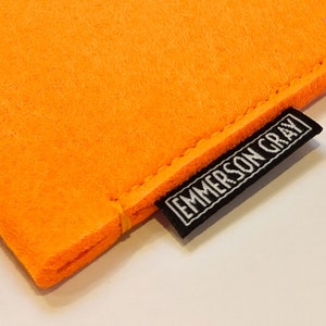 May include: Close-up of a bright orange felt pouch with a black label. The label, stitched to the pouch, reads "EMMERSON GRAY" in white capital letters. The pouch has a simple, clean design.