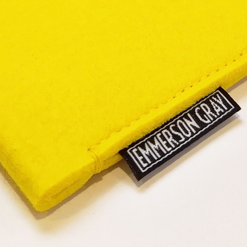 May include: Bright yellow felt pouch with a black and white label that reads "EMMERSON GRAY". The pouch is made of a soft, textured material and has a stitched seam. The label is rectangular and sewn onto the corner of the pouch.