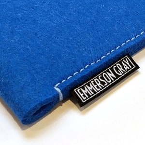 May include: Close-up of a bright blue felt sleeve with white stitching. A black label with the text "EMMERSON GRAY" in white is sewn onto the sleeve. The sleeve is made of a soft, textured material.