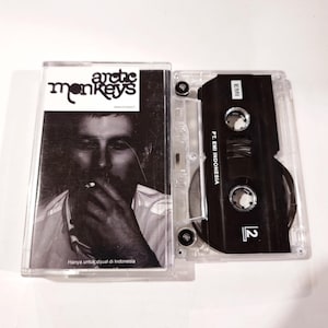 Könnte beinhalten: Eine Vintage-Kassette der Band Arctic Monkeys. Die transparente Hülle zeigt ein Schwarz-Weiß-Bild einer rauchenden Person. Der Schriftzug "Arctic Monkeys" befindet sich oben. Der Text "Hanya untuk dijual di Indonesia" steht unten.