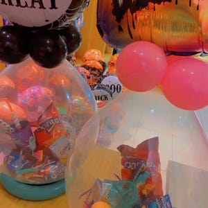 Puede incluir: Decoraciones de globos de Halloween. Un globo dice "Trick or Treat" con un diseño de telaraña. Otro dice "Happy Halloween". Globos transparentes llenos de caramelos y snacks, con globos rosas y negros.