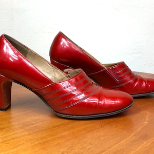 Puede incluir: Un par de zapatos de tacón de charol rojo con acabado brillante, punta redondeada, tacón de bloque y tres líneas horizontales cosidas en el empeine. El forro interior es de color beige claro y las suelas son de color marrón oscuro.