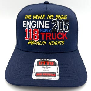 May include: A navy blue baseball cap with embroidered text. The text includes "FIRE UNDER THE BRIDGE", "ENGINE 205", "118 TRUCK", and "BROOKLYN HEIGHTS". A white sticker on the brim reads "OTTO CLASSIC FIT".