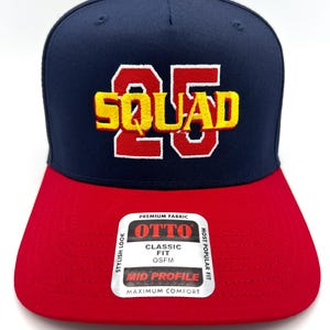 May include: A navy blue and red baseball cap with the word "SQUAD" and the number "25" embroidered in yellow and red. The cap has a flat brim and a red button on top. The label says "OTTO CLASSIC FIT OSFM".