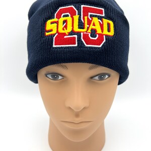 May include: Navy blue knit beanie with the word "SQUAD" and the number "25" embroidered in red and yellow. The beanie is displayed on a mannequin head against a white background.