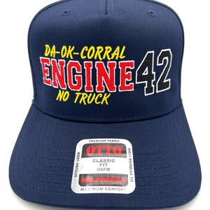 May include: A navy blue baseball cap featuring embroidered text that reads DA-OK-CORRAL ENGINE 42 NO TRUCK. The cap includes a sticker on the brim with the text OTTO PREMIUM FABRIC CLASSIC FIT OSFM MID PROFILE MAXIMUM COMFORT STYLISH LOOK.