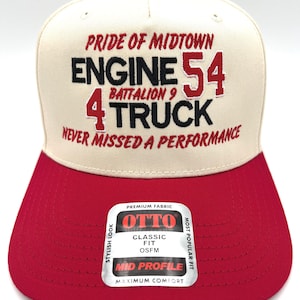 May include: A red and cream-colored baseball cap with the text "PRIDE OF MIDTOWN ENGINE 54 BATTALION 9 4 TRUCK NEVER MISSED A PERFORMANCE." The cap has a red brim and a small red button on top. A white sticker with the brand name "OTTO" is on the brim.