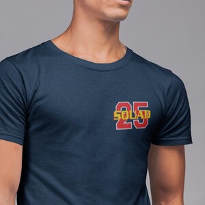 May include: A navy blue short-sleeved t-shirt featuring a graphic on the left chest. The graphic displays the number 25 in red with a white outline, with the word SQUAD written in yellow letters across the center of the number 25.
