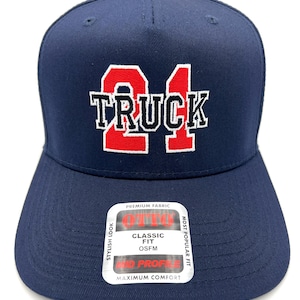 May include: A navy blue baseball cap featuring the red embroidered number 21 and the white embroidered text TRUCK centered on the front. A rectangular sticker on the brim displays the text OTTO, PREMIUM FABRIC, CLASSIC FIT, OSFM, MID PROFILE.