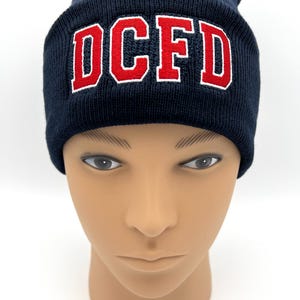 May include: Navy blue knit beanie with the embroidered letters "DCFD" in red with a white outline. The hat is displayed on a mannequin head against a white background. A warm winter hat.