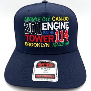 May include: A navy blue baseball cap with embroidered text in various colors. The text includes "EMERALD ISLE CAN-DO", "201 ENGINE", "TOWER 114", and "BROOKLYN TALLEY HO". A white tag with "OTTO" and "CLASSIC FIT" is on the front.