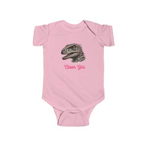 May include: Pink baby onesie with a graphic of a dinosaur wearing a pink flower headband. The text "Clever Girl" is printed below the dinosaur.