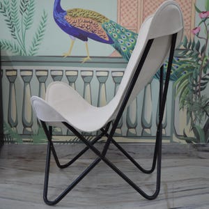 May include: A white butterfly chair with a black metal frame. The chair has a curved back and seat, and the frame is made of thin, angled metal rods. The chair is set against a wall with a peacock mural.