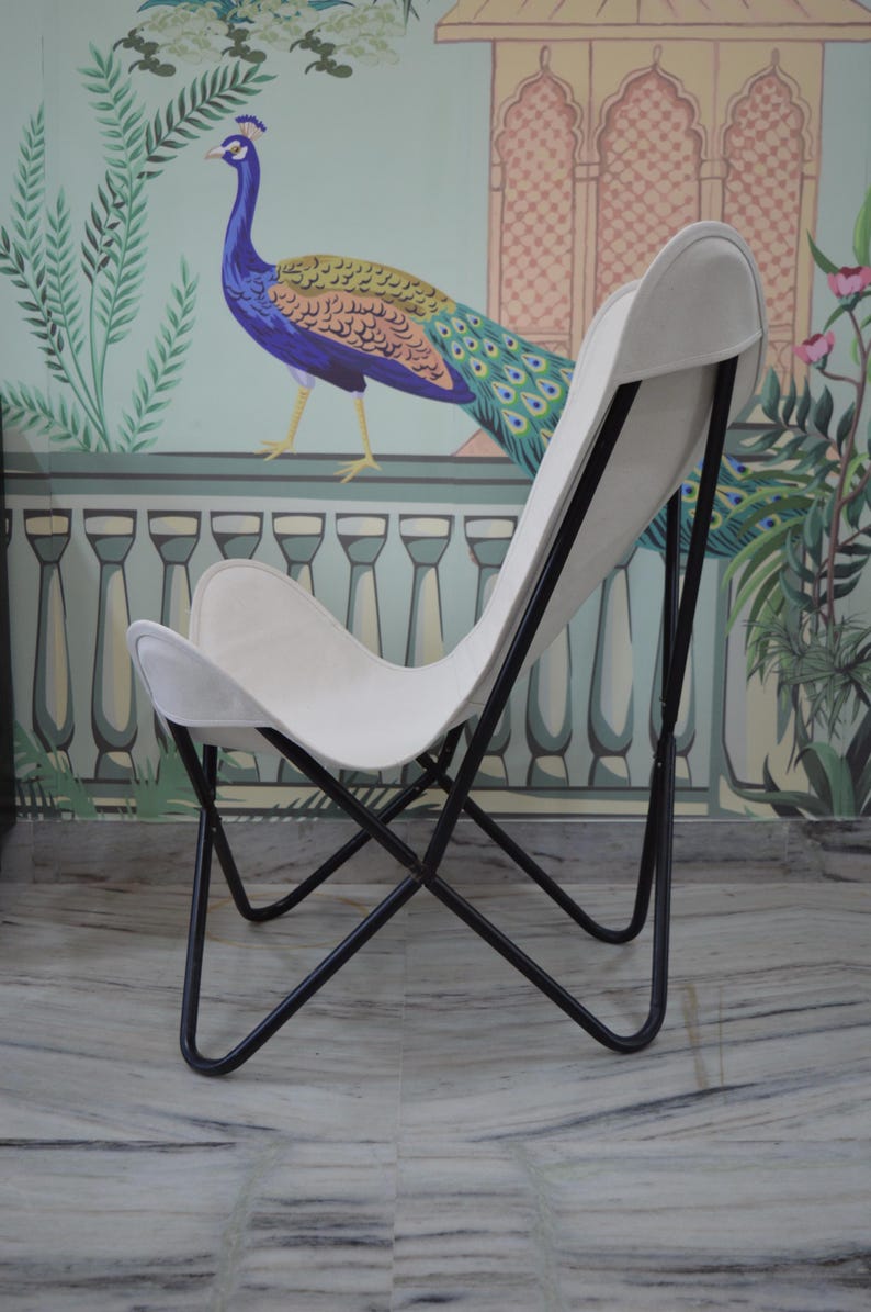 May include: A white butterfly chair with a black metal frame. The chair has a curved back and seat, and the frame is made of thin, angled metal rods. The chair is set against a wall with a peacock mural.