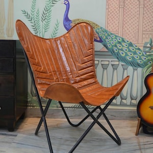 May include: A brown leather butterfly chair with a black metal frame. The chair is sitting on a white marble floor in front of a wall with a peacock design.