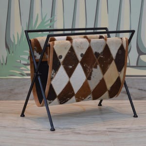 May include: A black metal magazine rack with a diamond patterned cowhide rug. The rug is brown, white, and black.