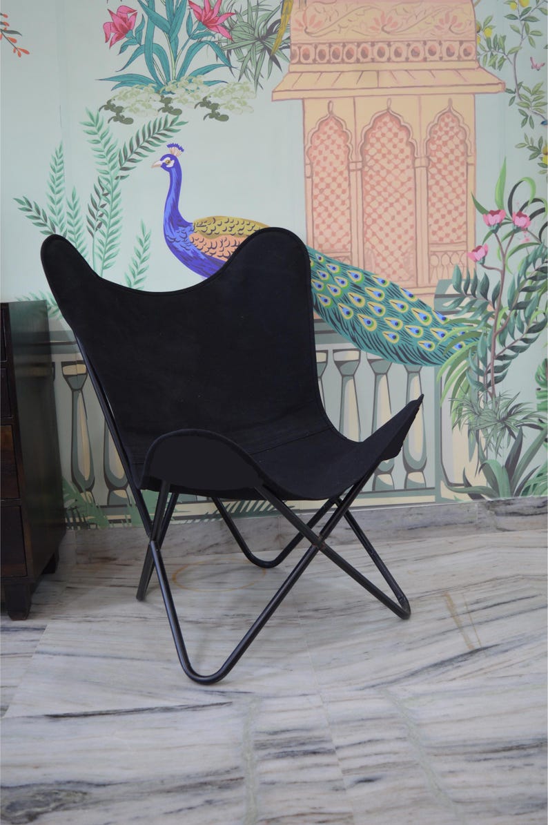 May include: Black butterfly chair with a metal frame. The chair has a distinctive, curved silhouette and a fabric seat. The chair is set against a backdrop of a colorful mural featuring a peacock and floral elements.