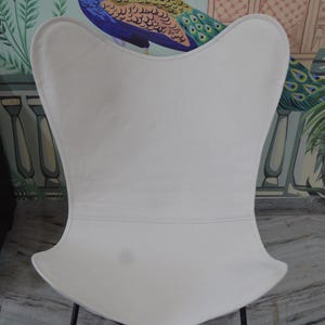 May include: A white butterfly chair with a black metal frame. The chair has a curved back and seat, and the frame is made of thin metal rods that cross at the base. The chair is set against a backdrop of a peacock mural.