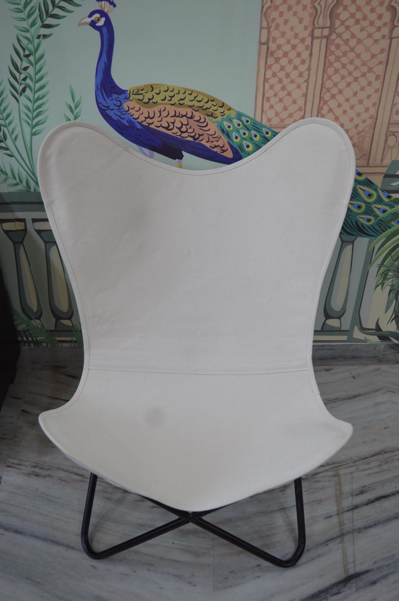 May include: A white butterfly chair with a black metal frame. The chair has a curved back and seat, and the frame is made of thin metal rods that cross at the base. The chair is set against a backdrop of a peacock mural.