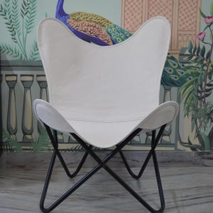 May include: White butterfly chair with a black metal frame. The chair has a curved seat and backrest, and the frame is made of intersecting metal bars. The chair is set against a wall with a peacock mural.