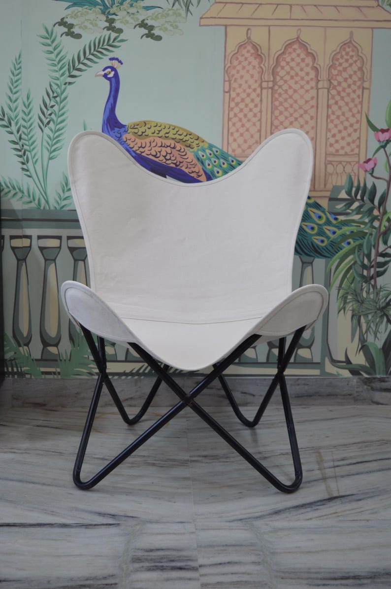 May include: White butterfly chair with a black metal frame. The chair has a curved seat and backrest, and the frame is made of intersecting metal bars. The chair is set against a wall with a peacock mural.