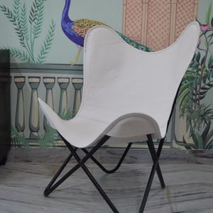 May include: White butterfly chair with a black metal frame. The chair has a distinctive wing-shaped design and is set against a backdrop of a peacock mural. The chair's seat is made of a smooth, solid white material.