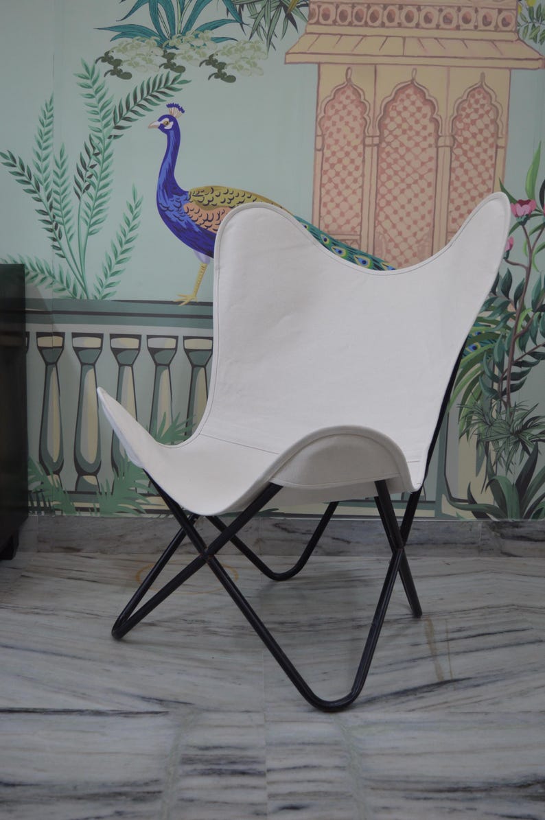 May include: White butterfly chair with a black metal frame. The chair has a distinctive wing-shaped design and is set against a backdrop of a peacock mural. The chair's seat is made of a smooth, solid white material.