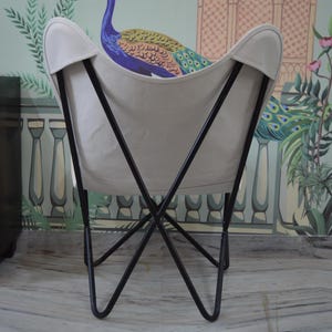 May include: A butterfly chair with a cream-colored canvas seat and a black metal frame. The chair is set against a backdrop of a wall mural featuring a peacock and floral design. The chair's design is reminiscent of a butterfly's wings.