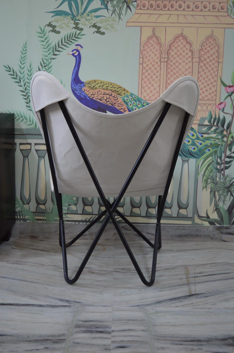 May include: A butterfly chair with a cream-colored canvas seat and a black metal frame. The chair is set against a backdrop of a wall mural featuring a peacock and floral design. The chair's design is reminiscent of a butterfly's wings.