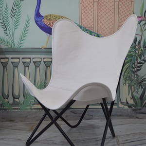 May include: White butterfly chair with a black metal frame. The chair has a distinctive curved shape and is set against a backdrop of a peacock mural. The chair's design is reminiscent of a butterfly's wings.