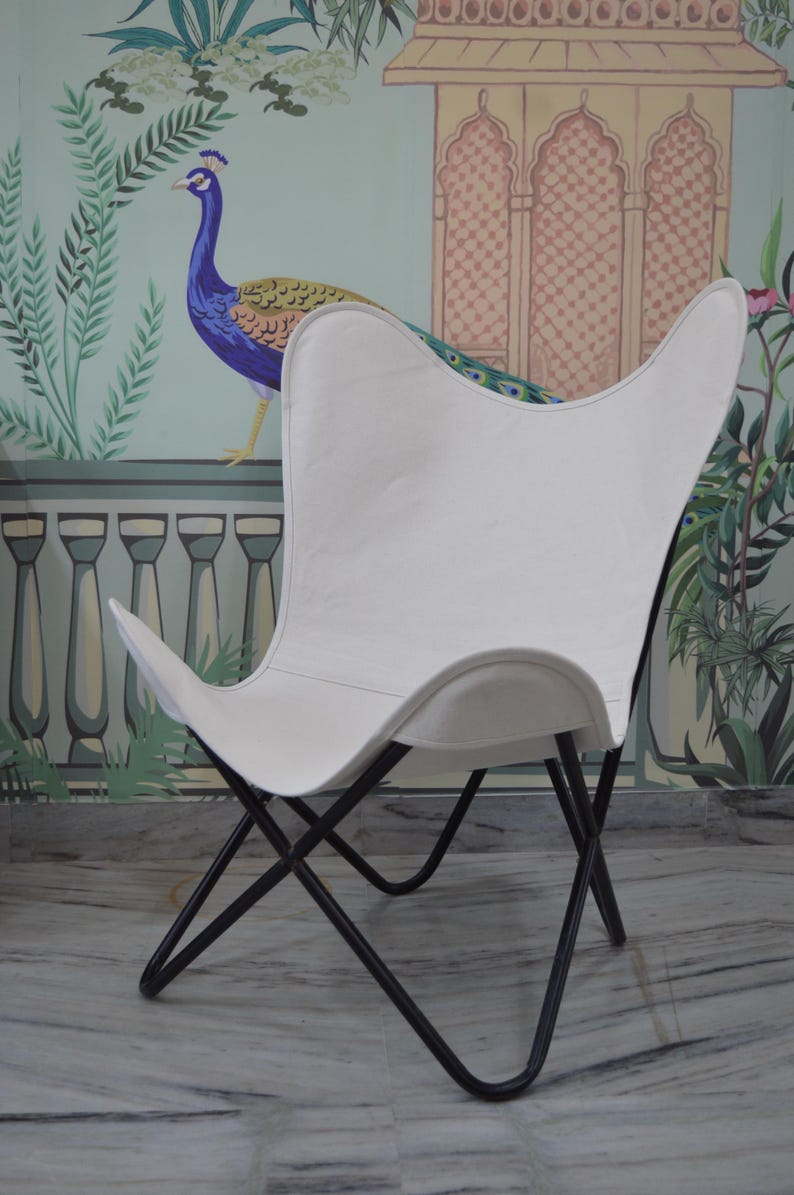 May include: White butterfly chair with a black metal frame. The chair has a distinctive curved shape and is set against a backdrop of a peacock mural. The chair's design is reminiscent of a butterfly's wings.