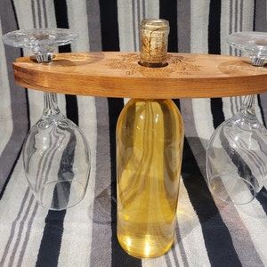 May include: Wooden wine bottle and glass holder with a decorative design. The holder has two slots for glasses and a center space for a bottle. The bottle is filled with a clear liquid.
