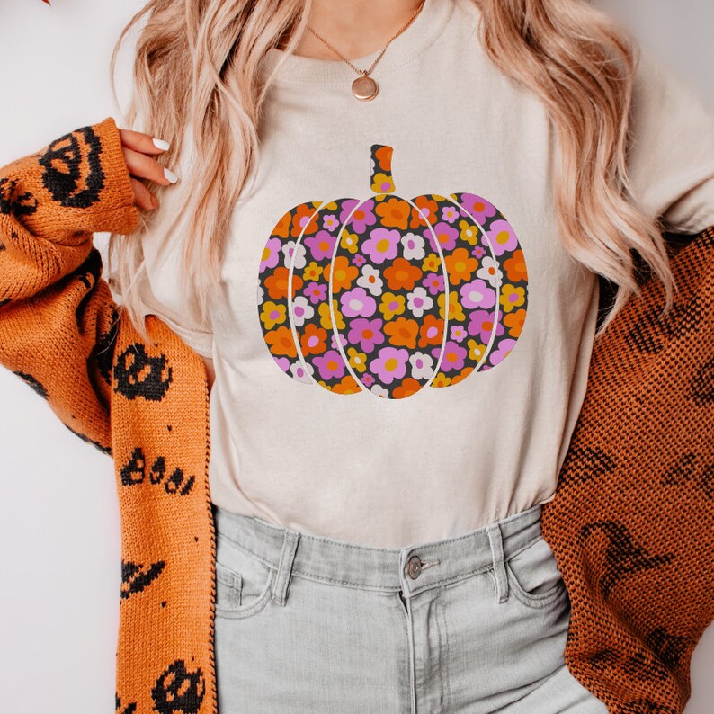May include: A white t-shirt with a floral pumpkin design. The pumpkin is orange, pink, and yellow with a black outline. The shirt is being worn by a person with long blonde hair.